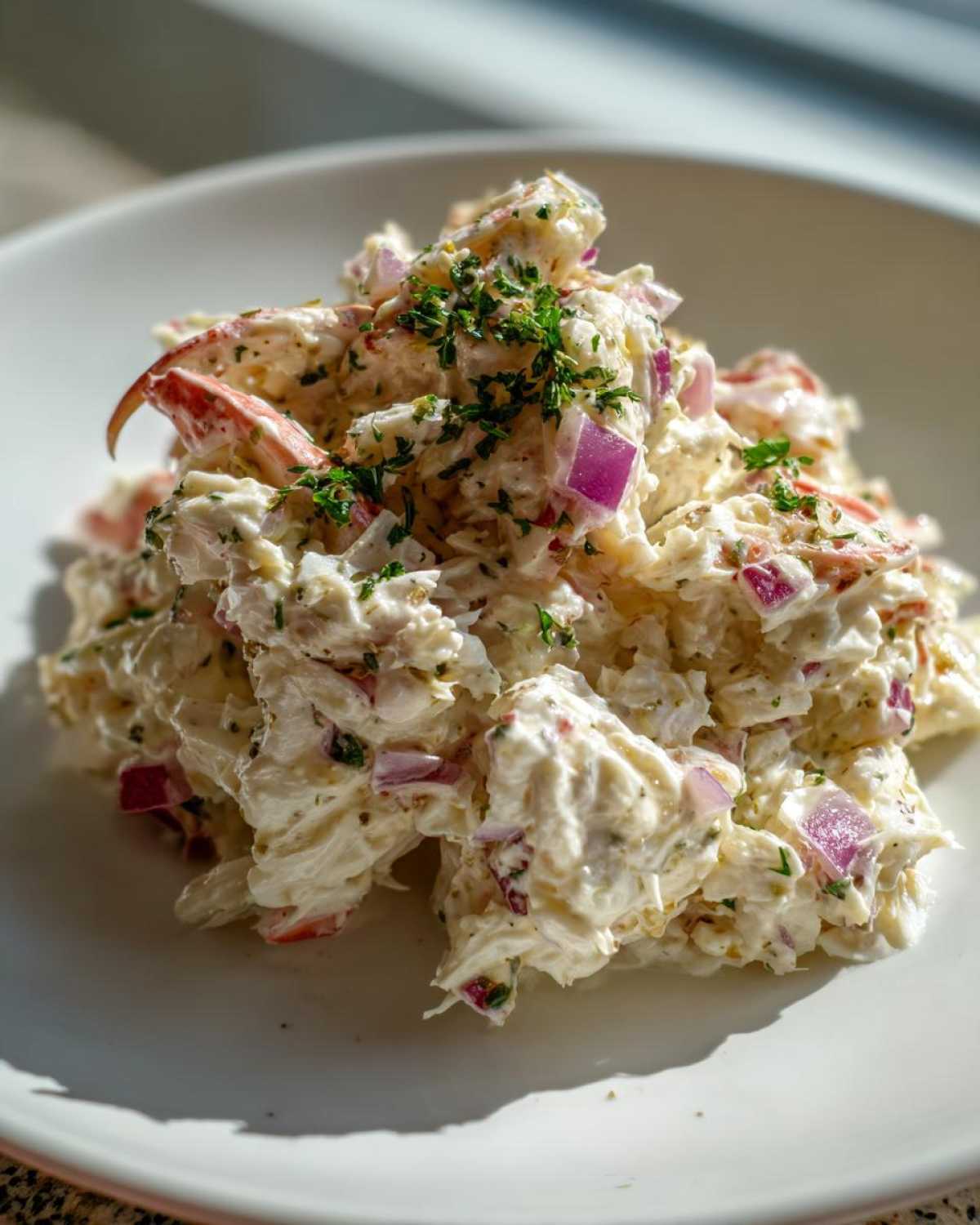 A mound of creamy crab salad mixed with diced red onion and topped with fresh parsley, served on a white plate.