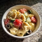Close-up of a white bowl filled with creamy Tortellini Pasta Salad featuring cherry tomatoes, cucumbers, and black olives.