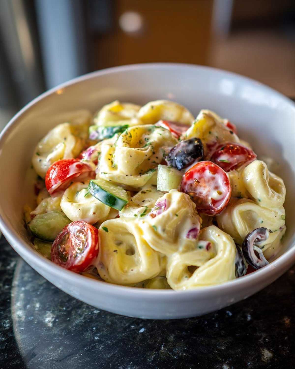 A close-up of creamy Tortellini Pasta Salad mixed with cherry tomatoes, cucumber, and black olives in a white bowl.