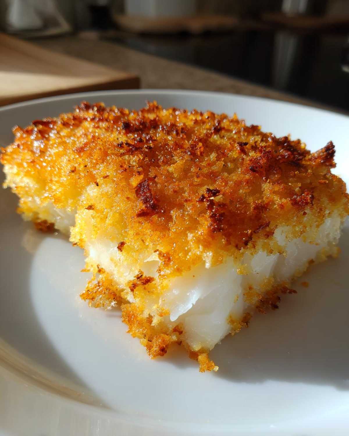 Close-up of a flaky white fish fillet with a golden, crispy baked cod topping on a white plate.