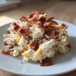 A mound of shredded Crockpot Crack Chicken (Bacon Cheddar Ranch) topped generously with crispy bacon pieces on a white plate.