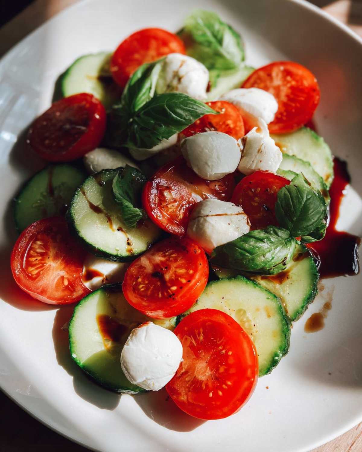 Close-up of a fresh Cucumber Caprese Salad featuring sliced cucumbers, tomatoes, mozzarella balls, and basil leaves drizzled with balsamic glaze.