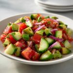 A vibrant bowl of fresh Cucumber Tomato Avocado Salad, featuring diced tomatoes, green cucumber, avocado chunks, and red onion.