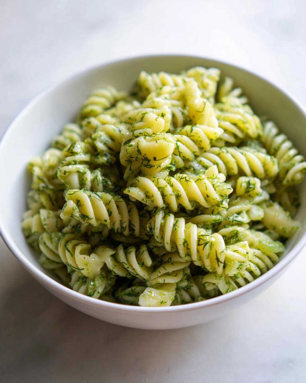 Close-up of fusilli pasta coated in a vibrant green dressing, featuring fresh dill, in a white bowl, showcasing the Dill Pickle Pasta Salad.