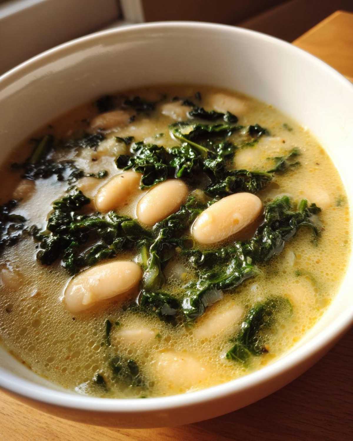 Close-up of a white bowl filled with Easy Tuscan White Bean Soup, featuring cannellini beans and dark green kale in a savory broth.