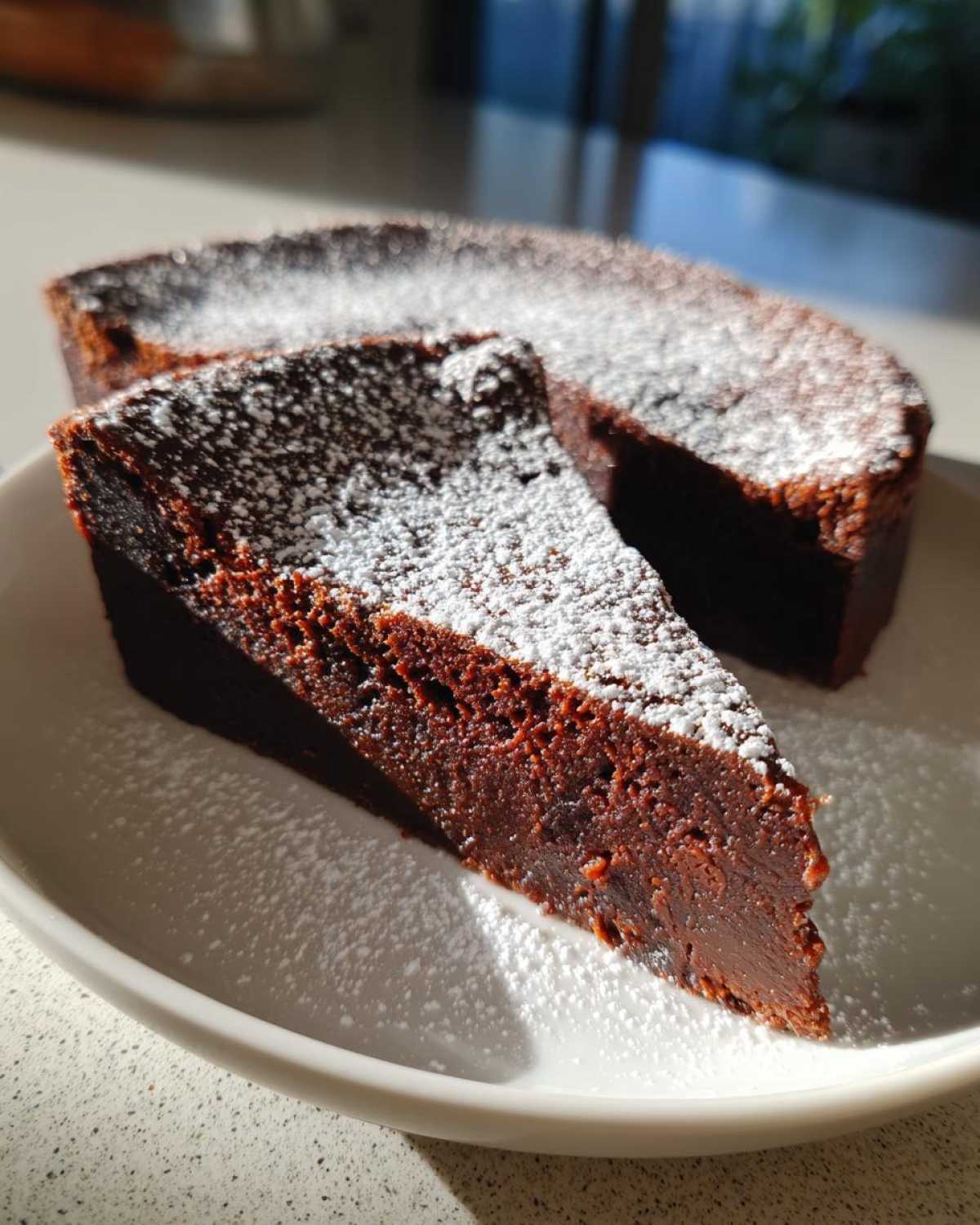 A decadent slice of flourless chocolate torte dusted heavily with powdered sugar, sitting next to the rest of the cake.