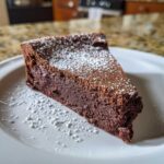 A decadent slice of flourless chocolate torte dusted with powdered sugar, resting on a white plate.