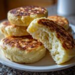 Close-up of homemade english muffins stacked on a plate, one broken open revealing a fluffy, airy interior.