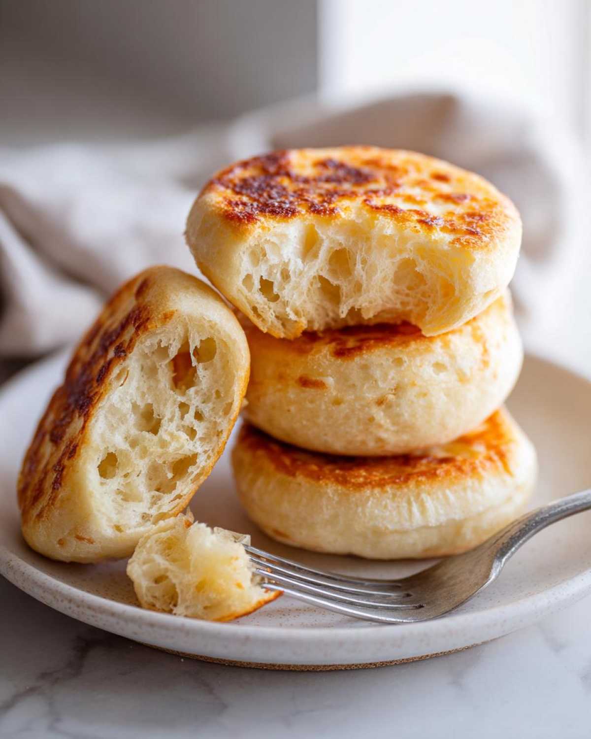 A stack of homemade english muffins, one split open showing the signature open, airy crumb structure.
