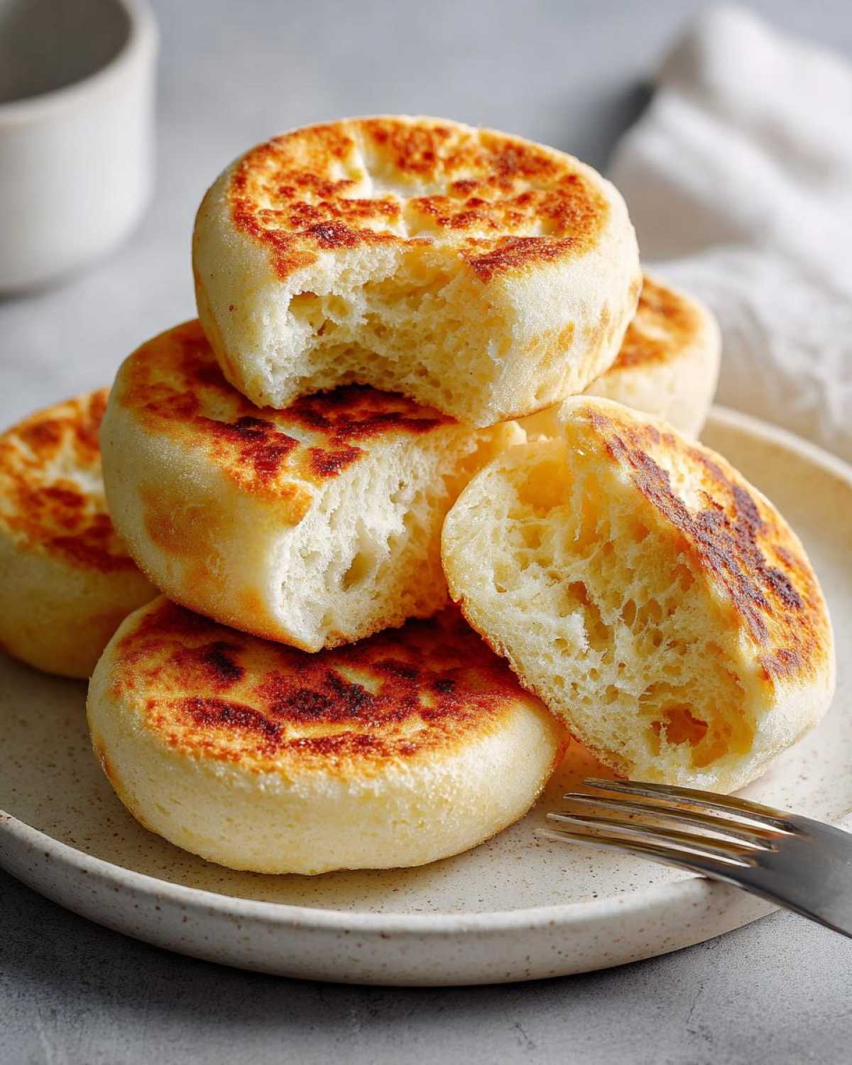 A stack of golden-brown homemade english muffins, one broken open to show the airy interior texture.