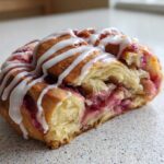 Close-up of a cross-section of a Geometric Rhubarb Cardamom Buns, showing flaky layers and a bright rhubarb filling, drizzled with white icing.