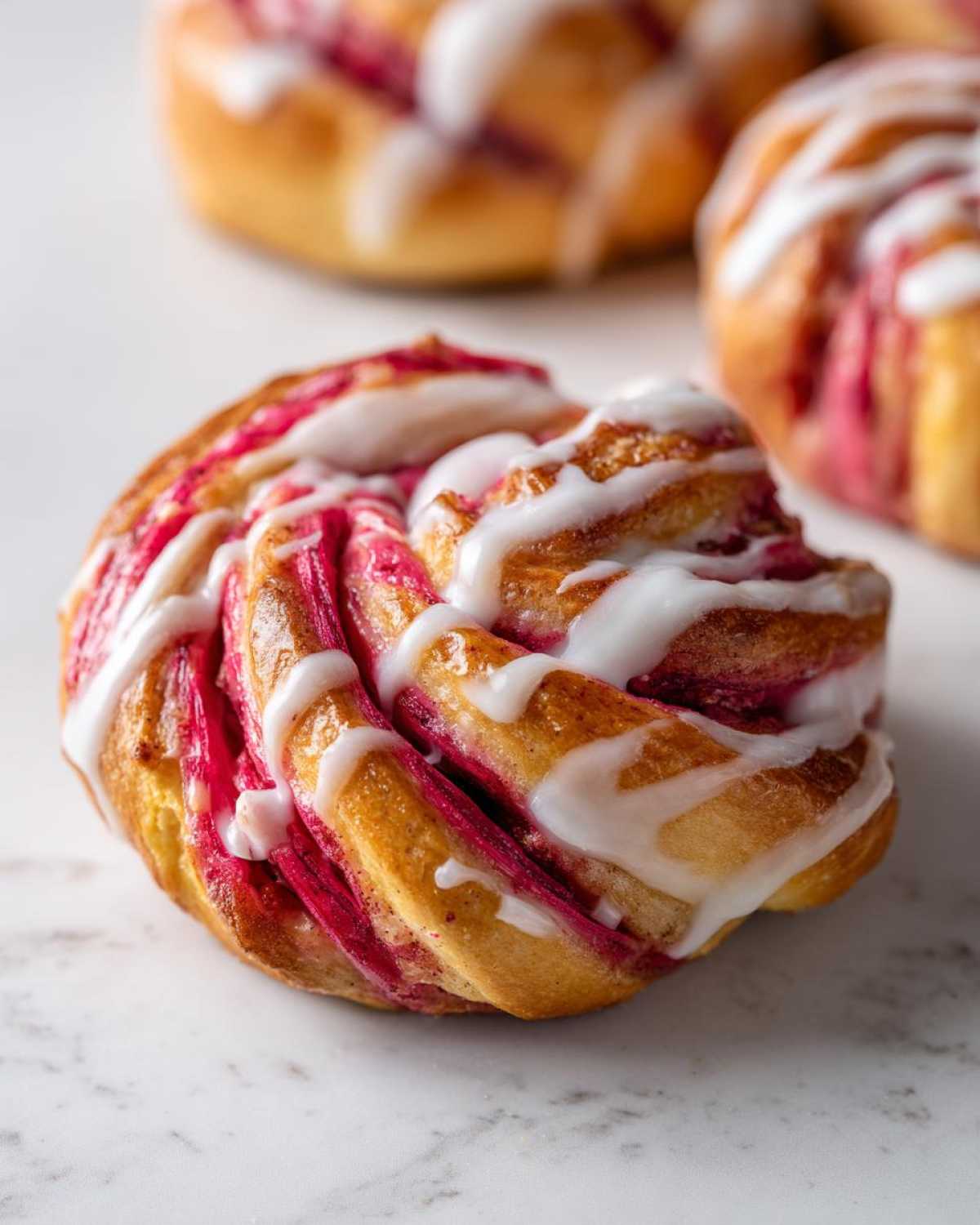 A close-up of a beautifully swirled Geometric Rhubarb Cardamom Bun, drizzled with white icing, resting on a marble surface.