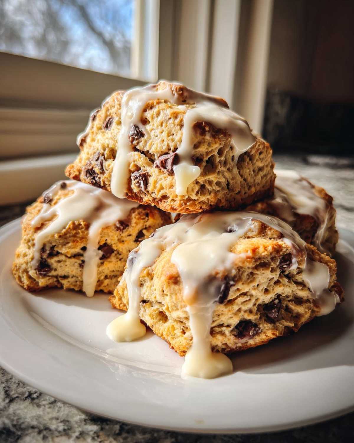 A stack of four freshly baked chocolate chip scones generously drizzled with white vanilla glaze, sitting on a white plate.