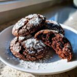 A plate of rich, dark Gluten-Free Chocolate Mint Cookies dusted heavily with powdered sugar, one cookie broken open to show the soft interior.