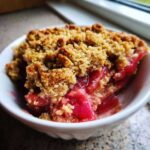 Close-up of a single serving of Gluten-Free Rhubarb Crisp showing bright pink rhubarb filling and golden oat topping in a white bowl.