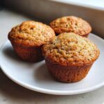 Close-up of three perfectly baked Vegan banana muffins with golden-brown tops resting on a white plate.