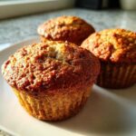 Three perfectly baked Vegan banana muffins with golden brown, textured tops sitting on a white plate.