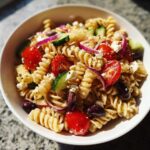 Close-up of a bowl filled with Greek Pasta Salad featuring rotini pasta, cherry tomatoes, cucumber, red onion, feta cheese, and Kalamata olives.