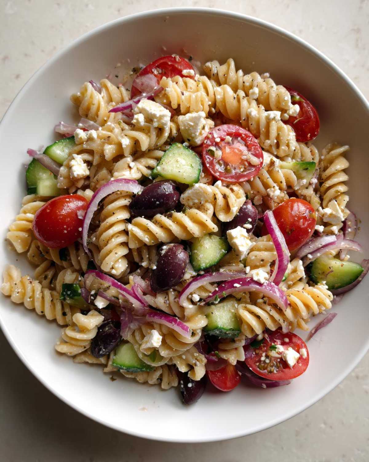 A close-up overhead view of a vibrant Greek Pasta Salad featuring rotini pasta, cherry tomatoes, cucumbers, red onion, olives, and feta cheese.
