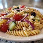A close-up serving of Greek Pasta Salad featuring rotini pasta, cherry tomatoes, cucumber, red onion, Kalamata olives, and feta cheese.