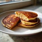 A stack of golden brown Healthy 3-Ingredient Banana Pancakes on a white plate, one pancake is broken open.