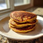 A stack of four golden-brown Healthy 3-Ingredient Banana Pancakes served on a white plate near a window.