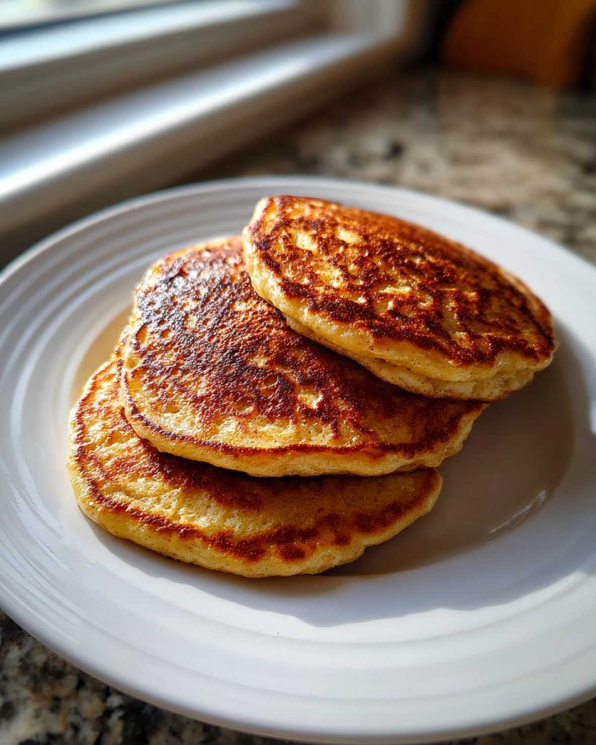 A stack of three golden-brown Healthy 3-Ingredient Banana Pancakes served on a white plate near a window.