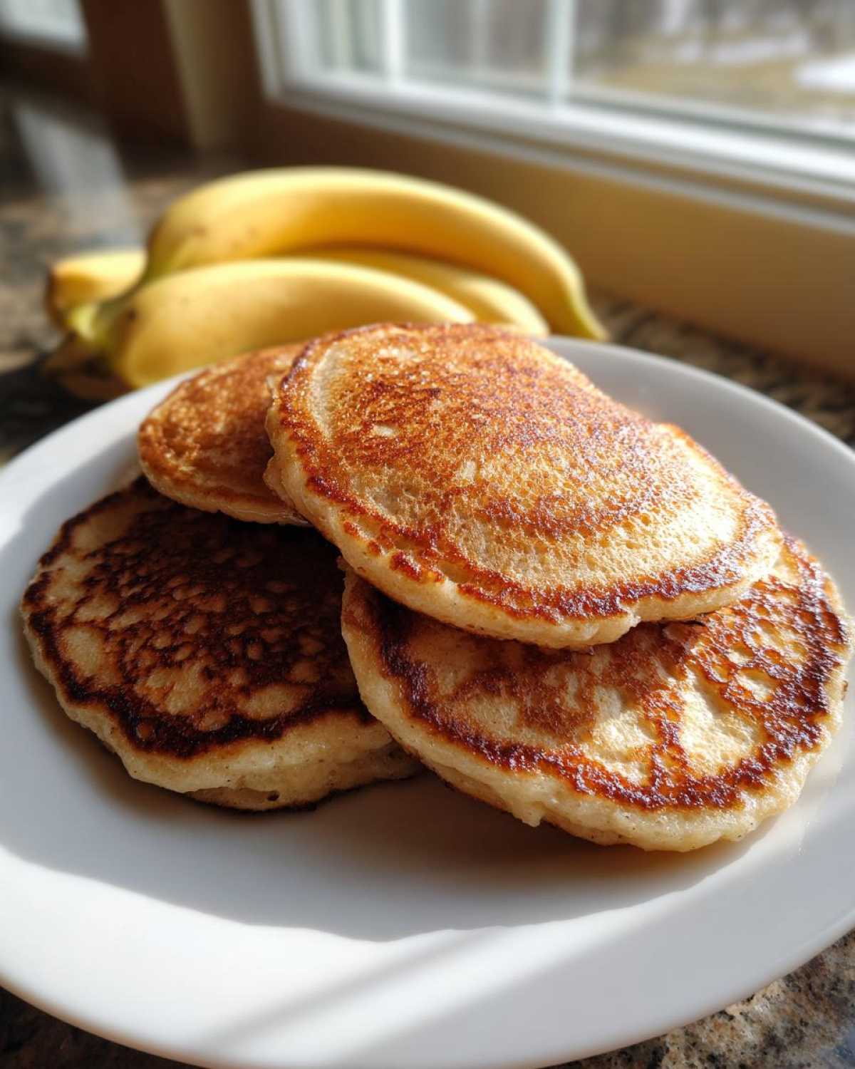 A stack of golden brown Healthy 3-Ingredient Banana Pancakes on a white plate with bananas in the background.