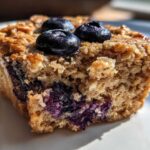 A close-up of a moist, golden-brown cottage cheese muffin filled with burst blueberries and topped with three fresh blueberries.