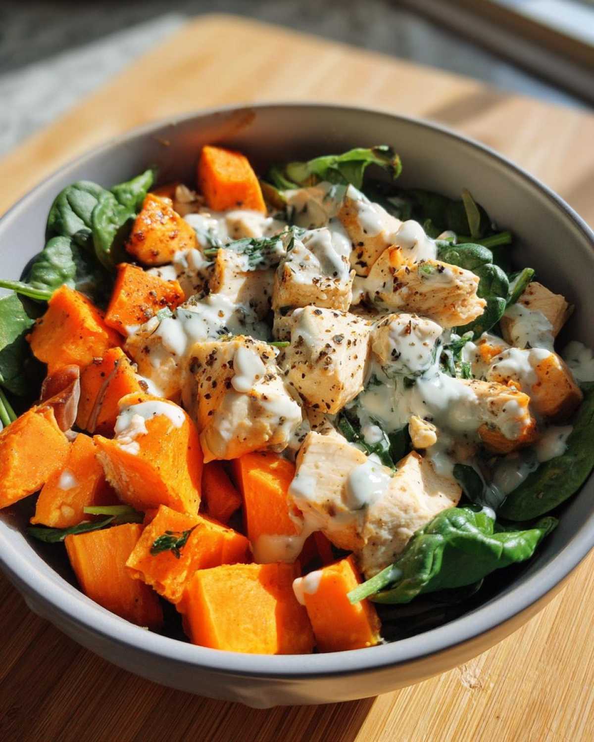 Close-up of a bowl containing High Protein Sweet Potato Chicken Bowls with chunks of chicken, roasted sweet potato, and spinach, drizzled with a white sauce.