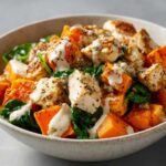 Close-up of diced chicken and roasted sweet potatoes mixed with spinach and drizzled with a creamy white sauce in a bowl, featuring High Protein Sweet Potato Chicken Bowls.