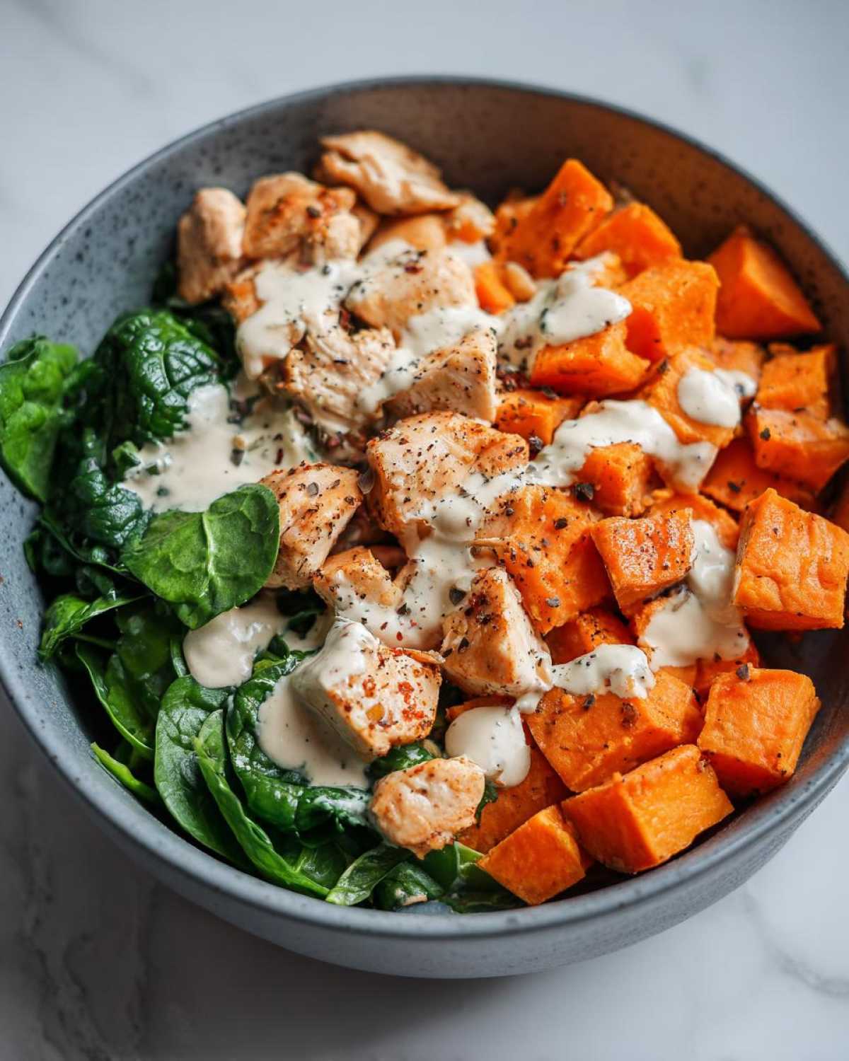 A close-up of a bowl containing High Protein Sweet Potato Chicken Bowls with spinach, diced chicken, and roasted sweet potatoes drizzled with a creamy sauce.
