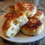 A stack of golden-brown, griddle-cooked english muffins on a white plate, one split open showing the airy interior.