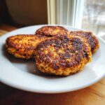 Four perfectly seared Homemade Salmon Patties stacked slightly on a white plate near a window.