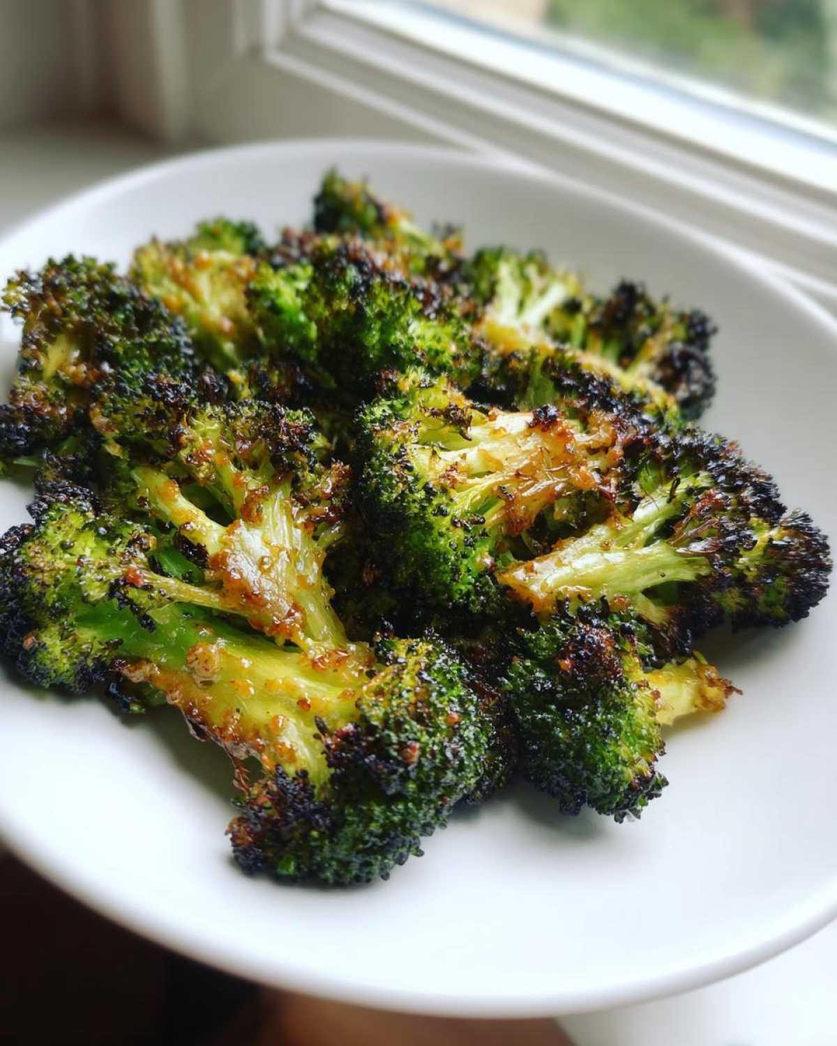 Close-up of perfectly charred Honey Mustard Roasted Broccoli florets served on a white plate near a window.