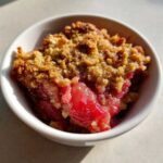 Close-up of a single serving of Irresistible Rhubarb Crisp with bright red filling and golden brown topping in a white bowl.