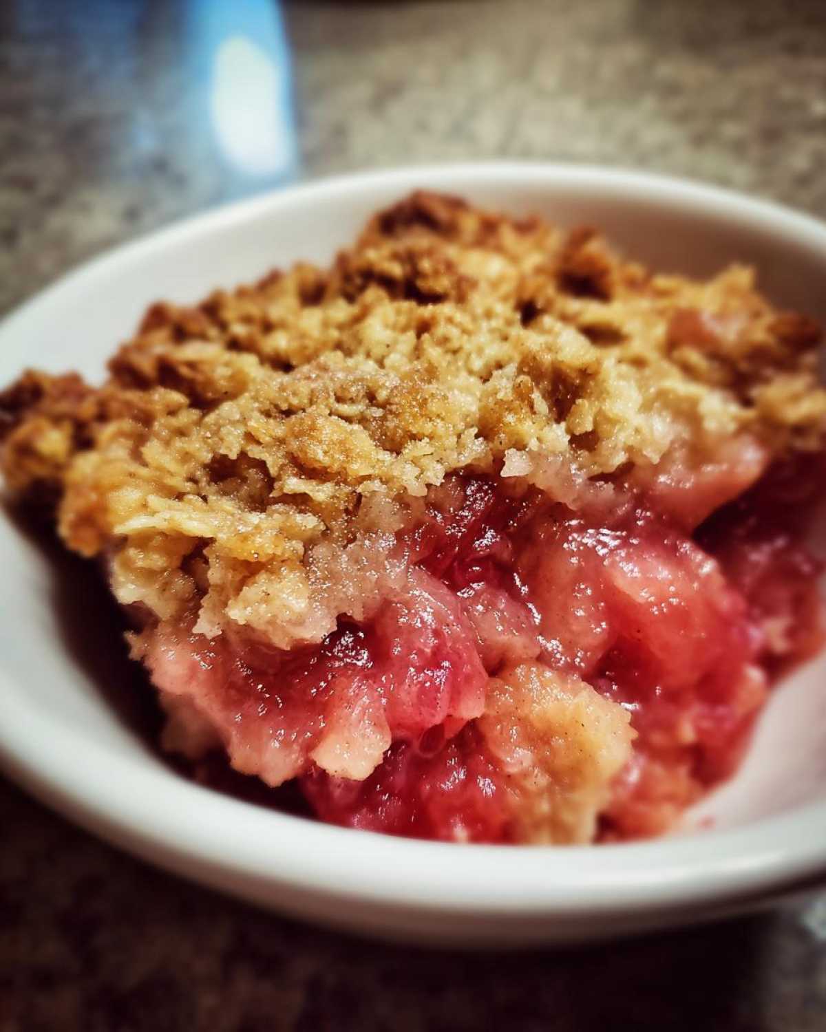 Close-up of a serving of Irresistible Rhubarb Crisp Recipe in a white bowl, showing juicy pink rhubarb filling and crunchy oat topping.