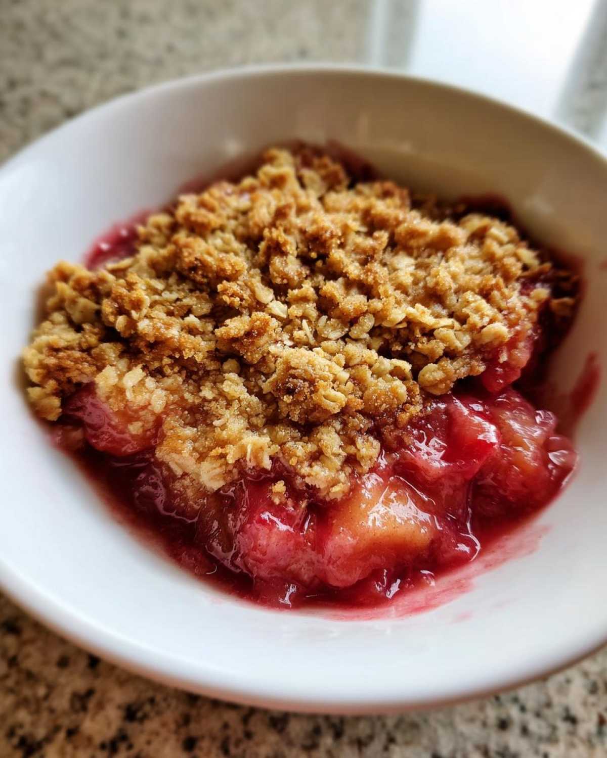 A close-up of a serving of Irresistible Rhubarb Crisp Recipe in a white bowl, showing baked oats and bright red fruit filling.