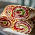 A stack of freshly cut italian pinwheels showing layers of deli meat, cheese, and lettuce rolled in a tortilla.