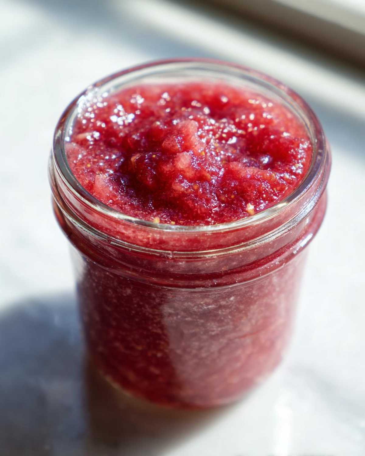 Close-up of a glass jar filled with bright red Rhubarb and Elderflower Jam (Pectin-Free) catching the sunlight.