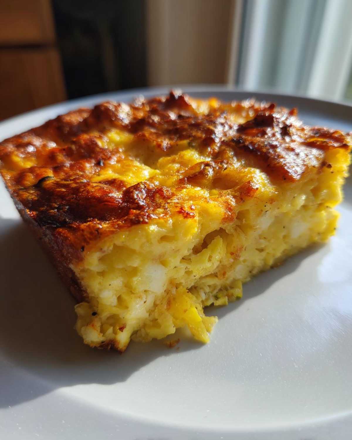 Close-up of a golden-brown slice of Keto squash casserole served on a white plate, showing a cheesy, baked texture.