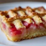 A close-up of a square slice of Lattice Rhubarb Snitter showing bright pink rhubarb filling under a golden lattice crust.