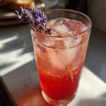 A refreshing, cold Lavender Rhubarb Spritzer garnished with fresh lavender sprigs, sitting in sunlight.