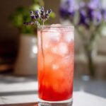 A tall, sweating glass filled with a vibrant pink Lavender Rhubarb Spritzer (Zero-Proof) over ice, garnished with fresh lavender.