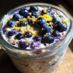 Close-up of a glass jar filled with Lemon Blueberry Overnight Oats, topped with fresh blueberries and bright yellow lemon zest.