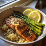 A close-up of a Lemon Herb Roasted Salmon & Asparagus Bowl featuring seasoned salmon over quinoa with roasted asparagus and lemon slices.