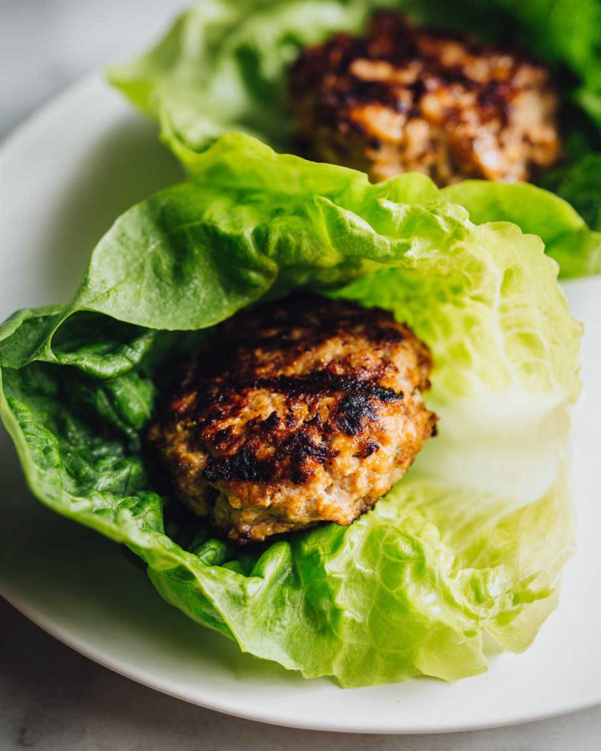 Two perfectly seared Lettuce Wrap Turkey Burgers nestled inside crisp green lettuce cups on a white plate.