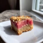 A close-up of one square of Low-Carb Rhubarb Dream Bars (Almond) with a crumbly topping and bright pink rhubarb filling.