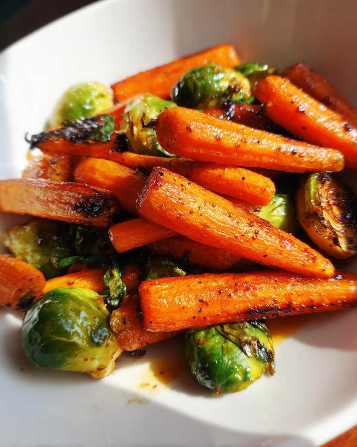 Close-up of roasted Maple Glazed Carrots and Brussels Sprouts coated in a shiny glaze in a white bowl.