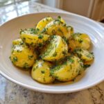 A white bowl filled with vibrant yellow, halved potatoes coated in a light dressing and fresh green herbs, ready to serve as Marinated Potato Salad.